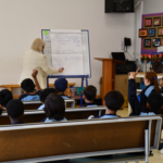 a classroom of school students and a teacher at the front writing on a white board